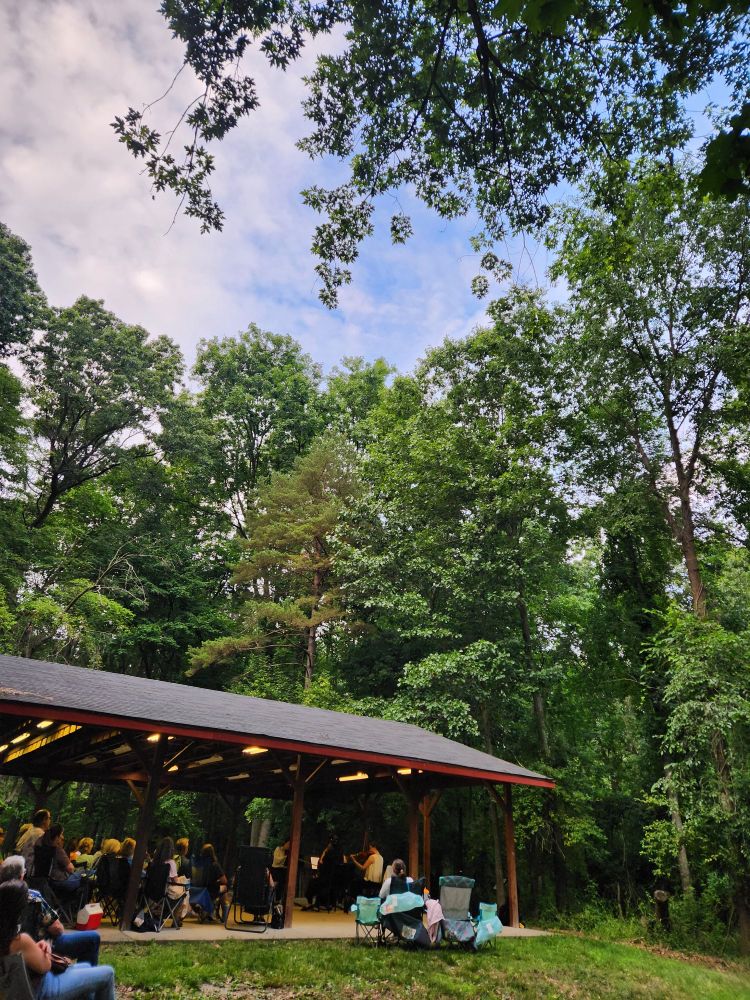 Outdoor string concert with people sitting under a pavilion in camping chairs in the midst of the trees