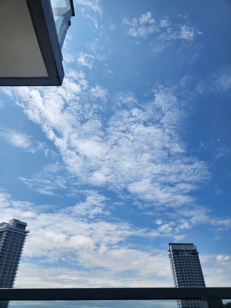 Clusters of thin white clouds in a blue sky. There's the edge of a balcony in the foreground on the top left side and a high-rise apartment building in the background on the bottom left and right sides