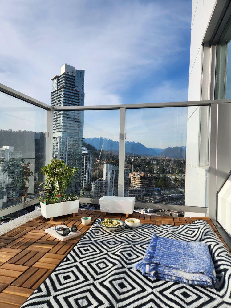 Photo of a high rise apartment balcony. There are wood tiles on the floor and a black and white mat on top. 