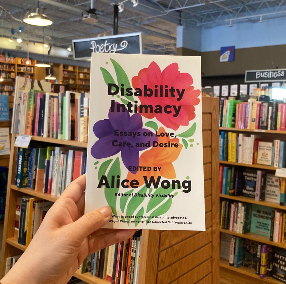 A hand holding up a copy of the book Disability Intimacy: Essays on Love, Care, and Desire, edited by Alice Wong, in a bookstore. In the background are shelves full of books and the poetry and business sections.