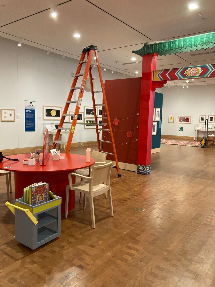 A museum exhibit almost ready for the public. In the foreground is a round red table with chairs and a toddler-sized dim sum cart with books on it. Beyond that is a ladder, and behind that is a replica of a Chinatown gate through which are blue-green walls with art on them. In the distance are the white walls of the gallery space with art on them.