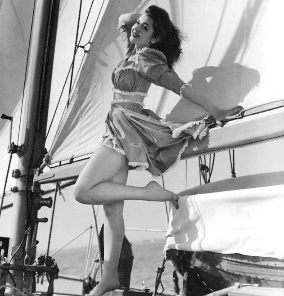 A black-and-white publicity photograph of actress Ann Rutherford, taken circa 1942, capturing the vivacity and poise characteristic of wartime Hollywood glamour. She poses playfully aboard a yacht, one leg bent and lifted, the other extended with pointed toes, her hand braced against the rigging while the other rests lightly atop her head in a gesture of carefree confidence. Her short, puff-sleeved dress, cinched at the waist and trimmed with delicate lace, flares in the breeze, revealing the clean, sculptural lines of her figure against the luminous sweep of the sails behind her. The interplay of sunlight and shadow lends the image a sense of sculptural clarity and motion, its composition evoking both nautical adventure and the exuberant optimism of early-1940s American femininity.