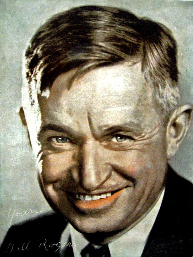 A hand-tinted studio portrait of a middle-aged man with neatly combed brown hair, wearing a dark suit and white shirt with a tie, smiling directly at the viewer. His expression conveys genial confidence, with bright eyes and a mischievous warmth suggesting both wit and approachability. The subtle tinting of his lips and skin gives the photograph an early 20th-century charm, bridging the line between photographic realism and painterly artifice. Inscribed faintly in cursive at the bottom left are the words “Yours, Will Rogers,” identifying the sitter as the beloved American humourist and social commentator. The portrait embodies the genial populism and understated sophistication of its era—an image at once intimate, idealised, and emblematic of a figure who made comedy a form of moral conscience.
