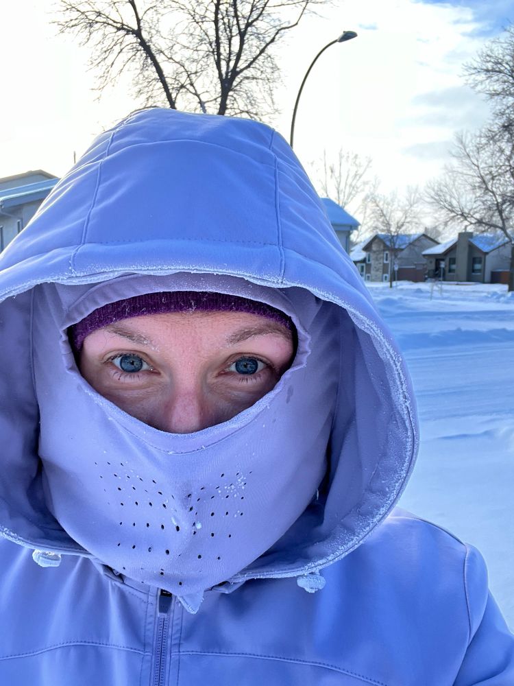 A woman is very bundled up in a light purple jacket, and face covering with only her eyes and bridge of her nose peeking out. Behind her is a lot of snow, and crisp winter morning light.