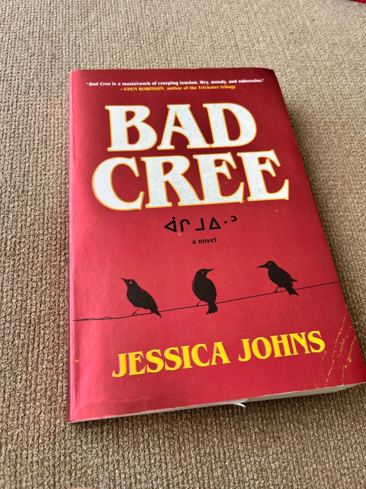 A paperback novel with a shiny red cover sits on a beige couch. The title, Bad Cree, is written in big white letters. Underneath in smaller black font are some Cree syllabics and the words “a novel”. A thin black line like a wire runs the width of the book near the bottom and three black crows sit on it. The author’s name, Jessica Johns, is in yellow under the crows. 