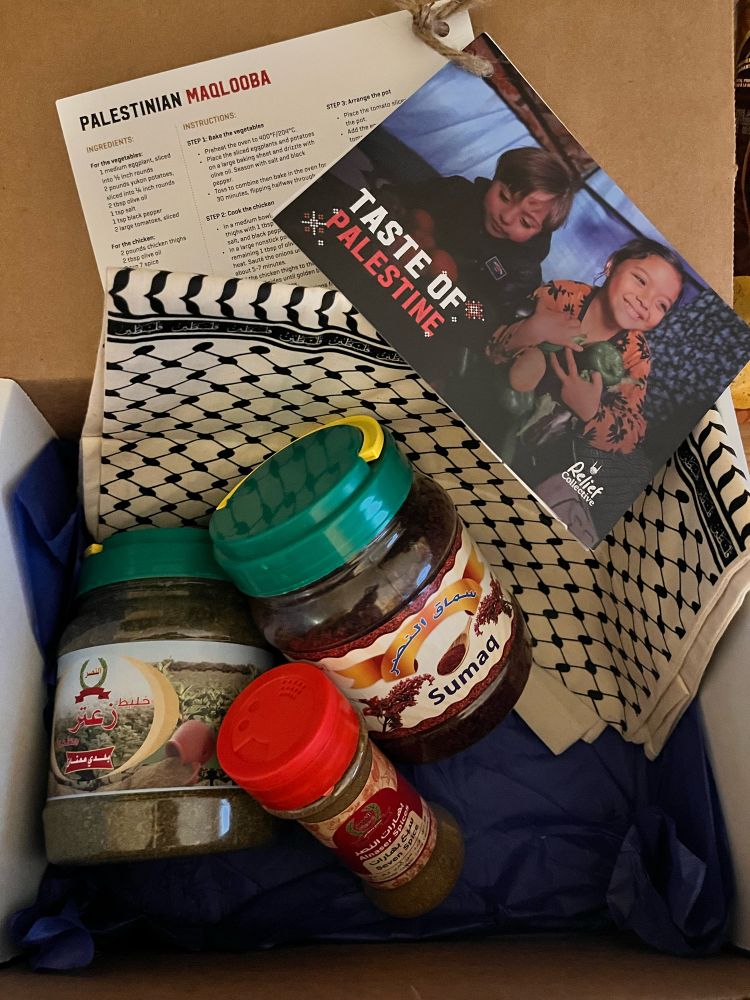 An open cardboard box shows two large jars with green lids, one labeled Sumaq, the other labeled in Arabic (but it is za’atar) and a smaller jar with a red lid labeled seven spice. There is a canvas tote bag folded up with keffiyeh designs, and some recipe cards labeled Taste of Palestine. 