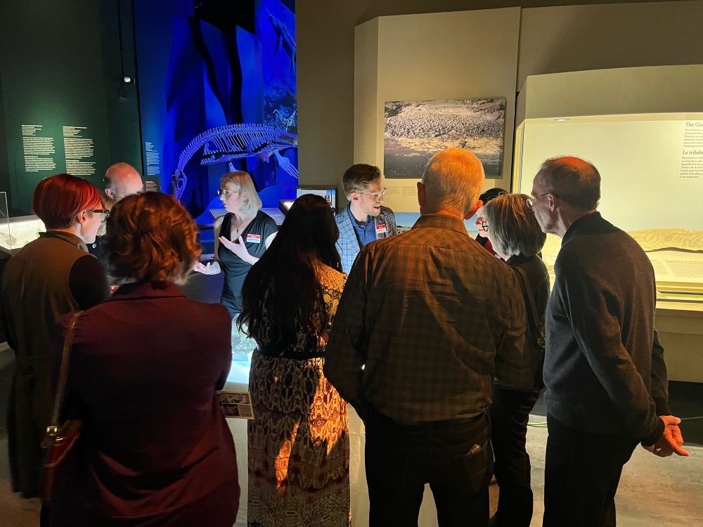 A group of people, many with their backs to the camera, chatting with two curators. The gallery lighting is dim, but the table in front of the curators with their specimens (unseen) is brightly lit, casting bright light on their faces. 