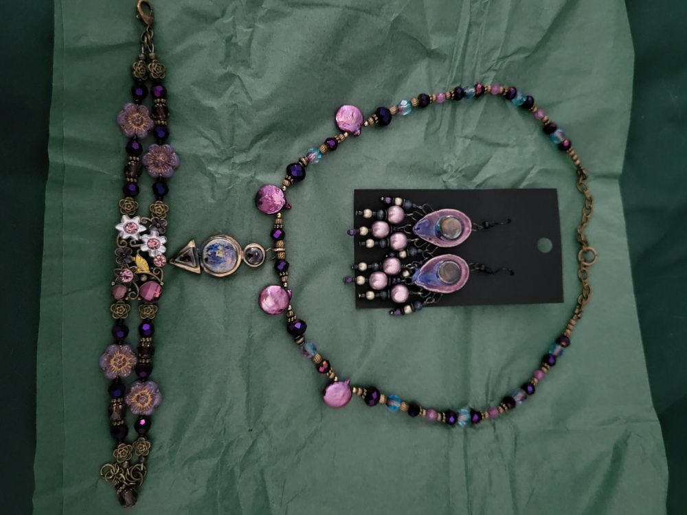 Earrings, a necklace, and a bracelet laid out on green tissue paper. All are in shades of purple and gold. The necklace also has some teal beads, while the bracelet has tiny flowers. Very summer-bleeding-into-fall, with light purples deepening into violet and then eggplant/aubergine.