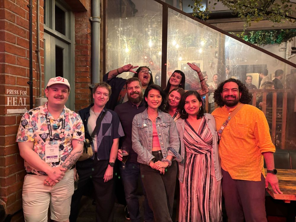 Group of people smiling at the camera. Two people are the back are doing fun poses. They are outside a venue, you can see people behind a screen in the background