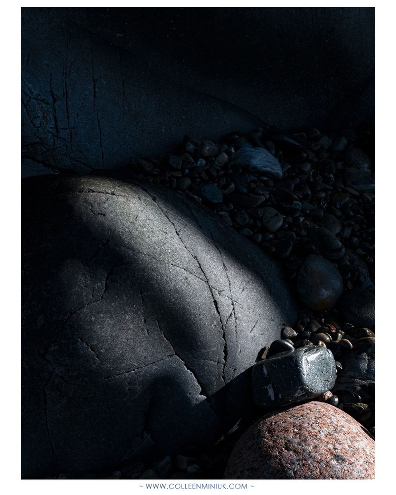 A grouping of different-sized coastal cobblestones made of granite and basalt are illuminated by sunlight as they rest against a shadowed background of cliff walls.