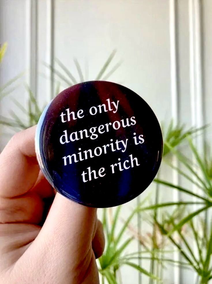 Meme of a Caucasian hand holding a black button that says “ the only dangerous minority is the rich” against a background of a white wall and some plants
