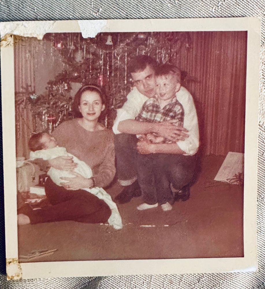 A small square battered sepia photo of two adults and two children posed in front of a Xmas tree 