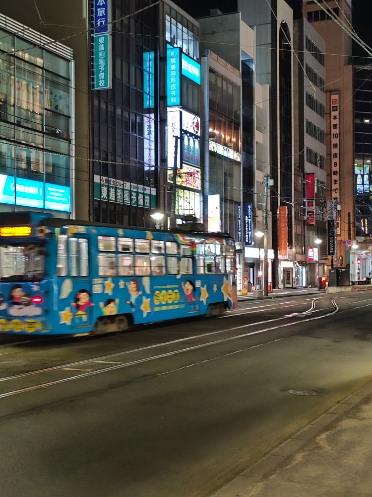 Sapporo Streetcar goes away from the photographer. The streetcar is blue with some drawings and it's seen from its right, from the sidewalk. It's a cityscape during the night.