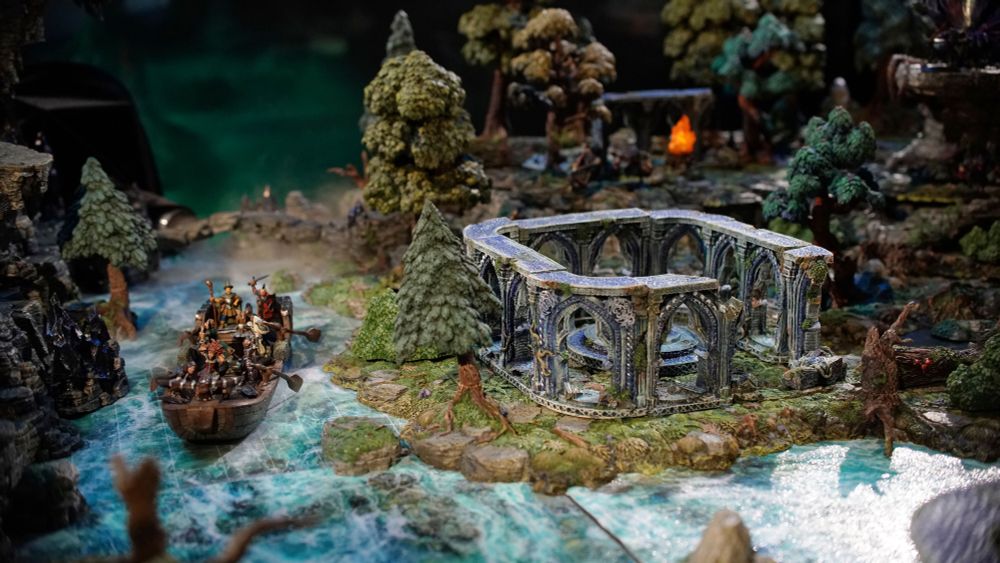 A miniature boat on a blue mat next to a gray elven ruin on a grassy land surrounded by trees.