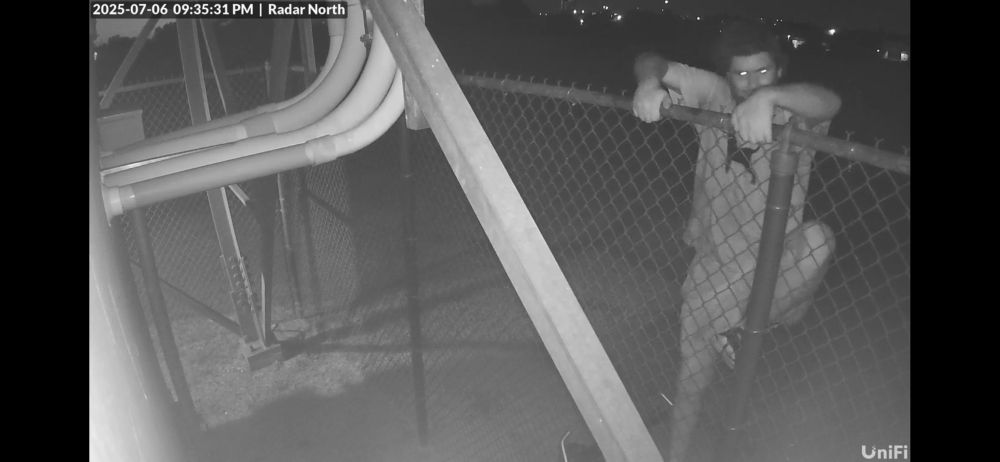 A frame of a black and white security camera video showing a man climbing over a chain link fence.