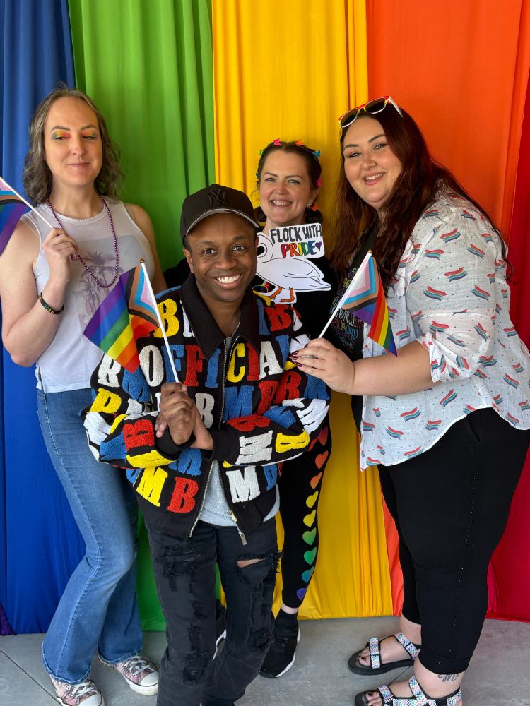 four people pose at the local pride event at the rainbow backdrop photo spot. They hold various props, primarily pride flags. o e person looks off to the left with a mischievous look, but I think she's just confused. There is no deviance afoot. Calm the fuck down