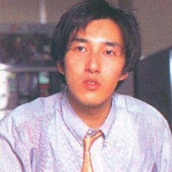 photo of goichi suda/suda51 from the 1996 twilight syndrome complete guidebook. he is around 28 years old here and in where i’m guessing is an office for human entertainment. he is wearing a blue dress shirt and a light orange tie. he is looking forwards and looks like he was in the middle of explaining something 