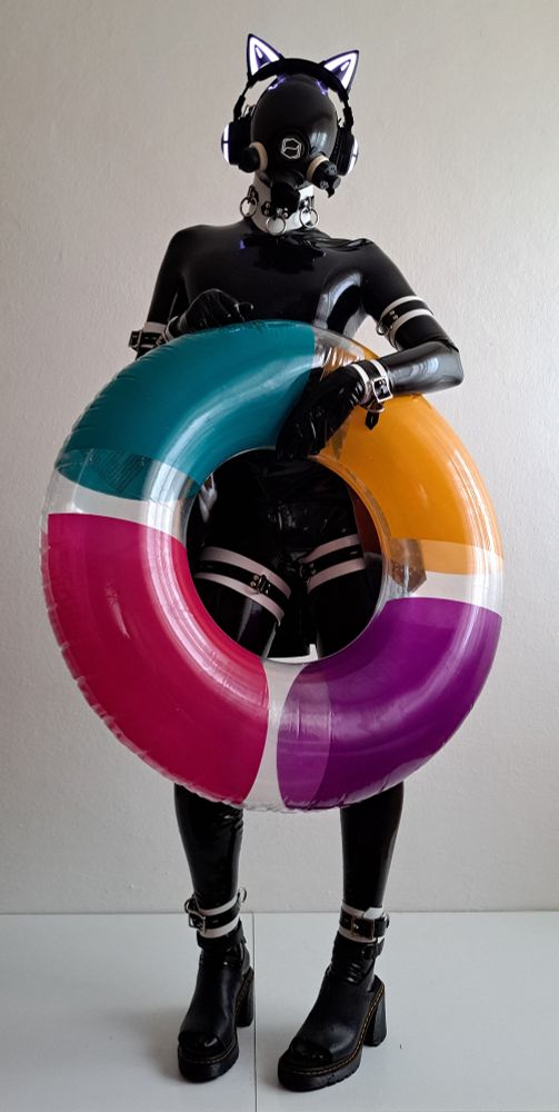 Photo of HexCorp drone 6919 holding a colored swim ring. It's wearing shiny black latex catsuit, waist corset, skirt, gloves, socks and DrMartens, mask and MSA Millennium gas mask with 3d printed attachments including HexCorp logo. All of that paired with black and white cuffs (ankle, thigh, wrist, arm and posture collar). The swim ring is conveniently lifting the skirt, revealing the thigh straps.