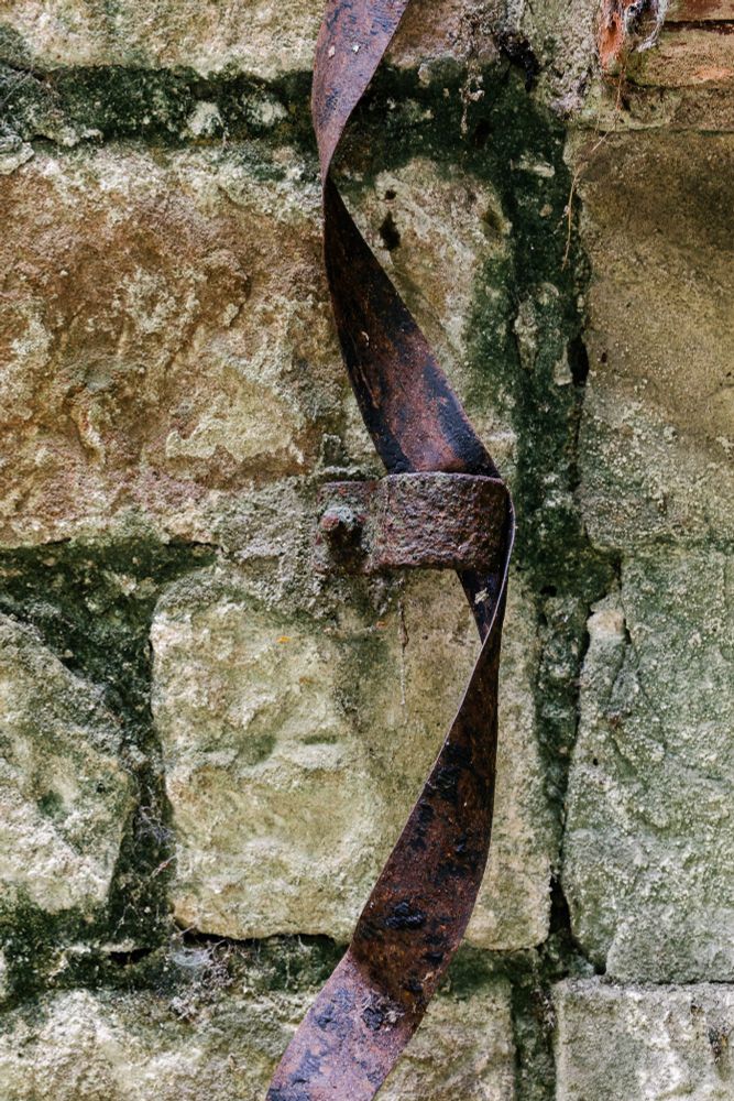 Das Bild zeigt eine grob gemauerte Steinwand mit unregelmäßigen, kantigen Steinen in erdigen Beige- und Grautönen. Zwischen den Steinen sind dunkle Fugen und moosgrüne Verfärbungen zu sehen, die auf Alter und Feuchtigkeit hindeuten. In der Mitte des Bildes verläuft ein rostiges, schmales Metallband, das sich in einer sanften Drehung nach unten windet. Es ist mit einem Bolzen an der Wand befestigt, und die Oberfläche des Metalls ist von Rostnarben, Kratzern und Abblätterungen gezeichnet. Das Bild vermittelt den Eindruck von Verfall und vergangener Handwerkskunst.