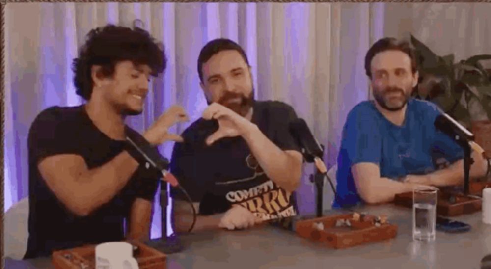 three men are sitting at a table with microphones and making a heart shape with their hands .