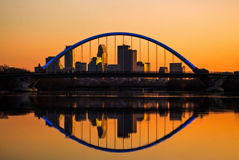Skyline of Minneapolis, framed by a suspension bridge, reflected in smooth water at sunset 