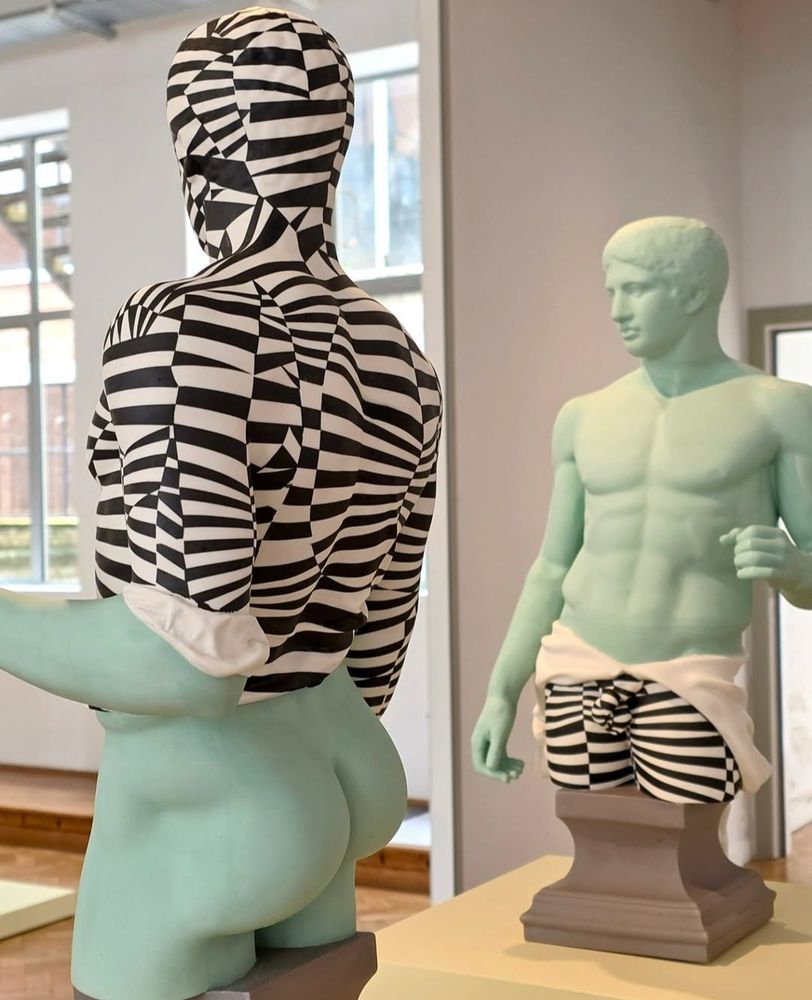 Two sculptures the shape of classical statues but a light teal colour. The bottom half of one is painted with black and white stripes, and the top half of the other is also