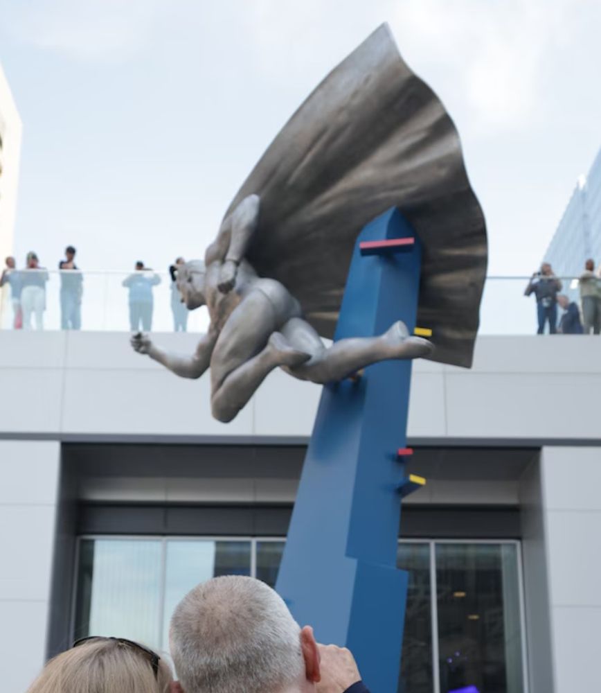 A photo showing two figures looking up at the dynamic sculpture of Superman in flight. With one knee bent and the other leg out straight, his contrapposto is in full evidence 