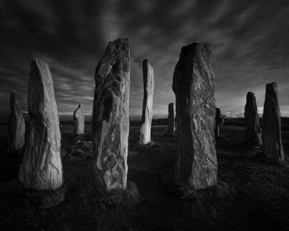 Stone Circle, Callanish is a landscape format photograph in black and white and shows a series of ancient standing stones. These megaliths vary in height and shape, some tall and slender, others shorter and more irregular. The surface of the stones is rough and textured, some with visible grooves or patterns eroded into them. The stones are situated on ground that appears grassy and uneven. The sky above the stones is streaky, with fast-moving clouds indicating a windy environment or a long exposure shot. There is a horizon in the distance, but no buildings or trees are visible and show a bare and rugged landscape.