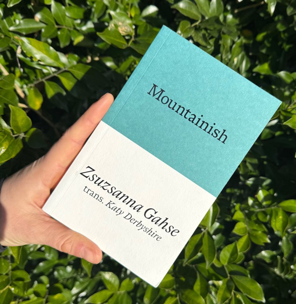 Me holding my copy of the book up in front of a leafy green hedge in the sunshine 