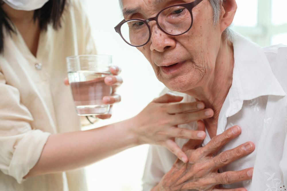 Eine ältere Person fasst sich mit der Hand an den Brustkorb. Eine zweite Person legt ihre Hand auf den Hals der ersten Person. In der anderen Hand hält sie ein Glas Wasser. 