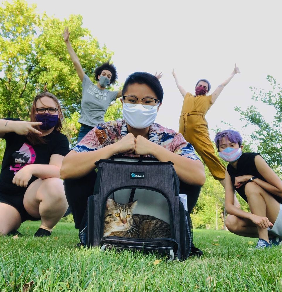 A photo of the Transplanar cast from 2020 posing outside in a park. Everyone is masked and posing around a backpack carrier with a tabby cat inside. The photo features: Erica, Lyra, Connie, Sea, and Max. 