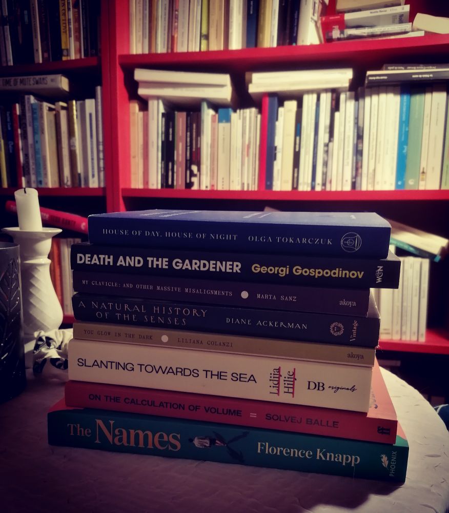 Book stack: Tokarczuk, House of Day House of Night; Gospodinov, Death of the Gardener; Sanz, My Clicle and other massive misalignments; Ackerman, A natural history of the senses; Colanzi, You glow in the dark; Hilje, Slanting towards the sea; Ball, Volume II; Knapp, the Names