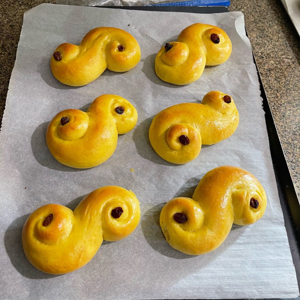 Six yellow buns on a tray of parchment paper, each one in an s-shape with a raisin in the center of each whirl