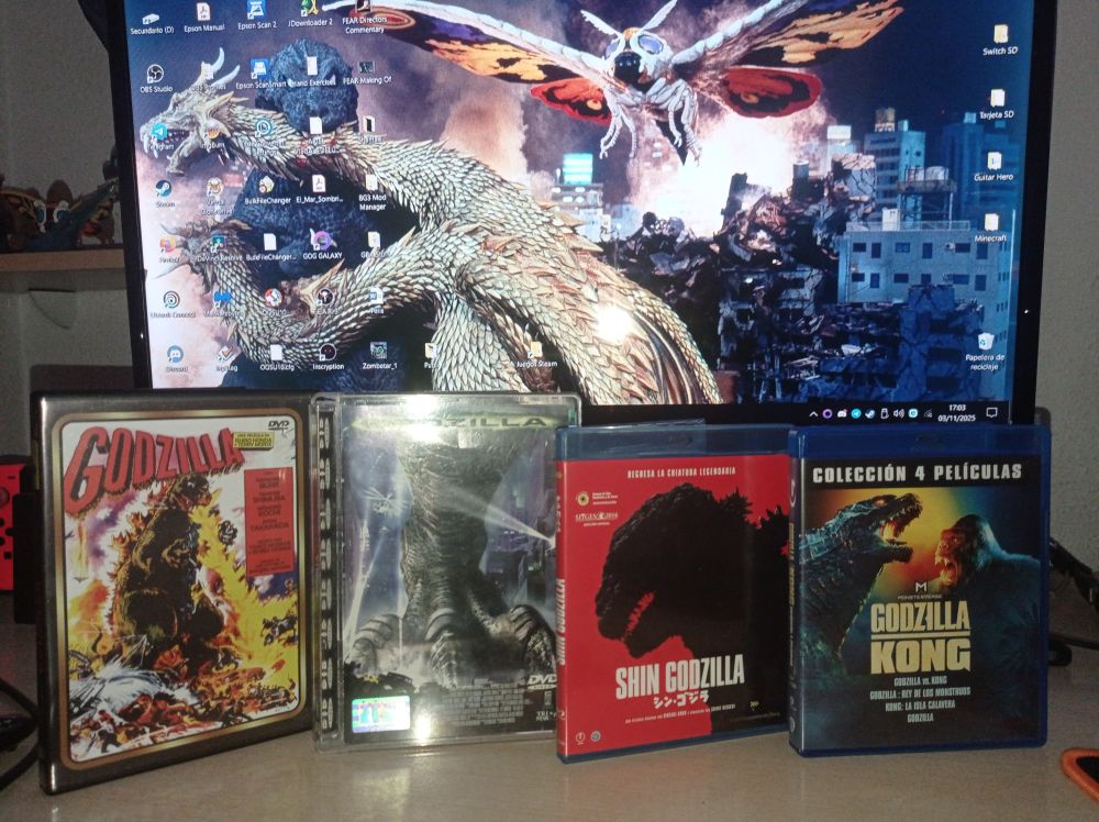 Photograph of various Godzilla movies (1954, 1998, Shin Godzilla and Monsterverse). Behind them is a PC screen with a shot of a movie with Godzilla, Ghidora and Mothra as a desktop wallpaper.