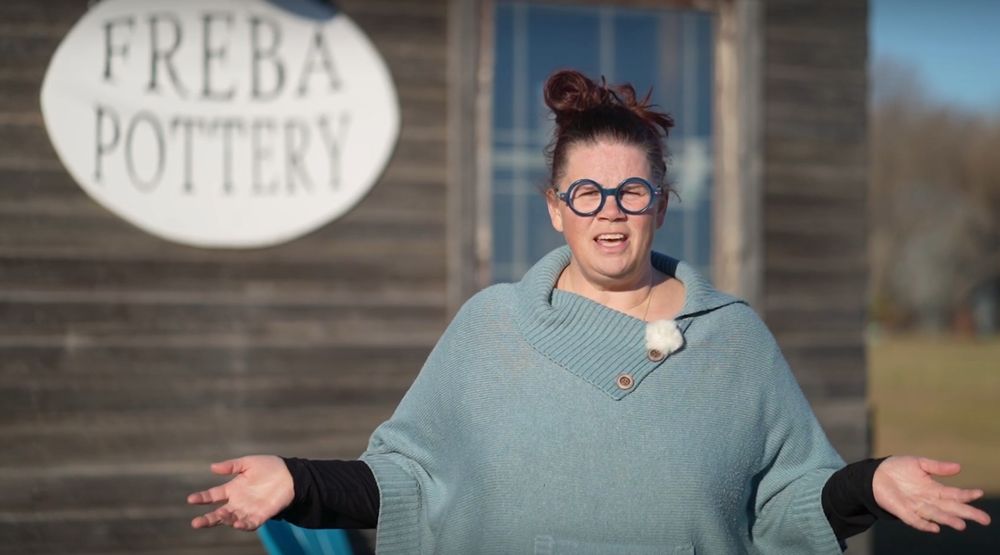 Woman with her hair up and glasses on, stands in front of a wooden pottery studio with her arms open. 