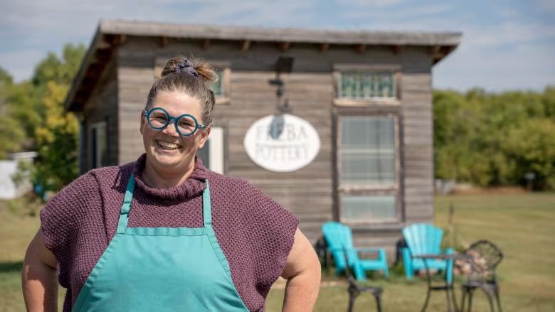 Woman with hair pulled back and funky blue glasses stands in front of a pottery shed. She wears an apron over her sweater. 