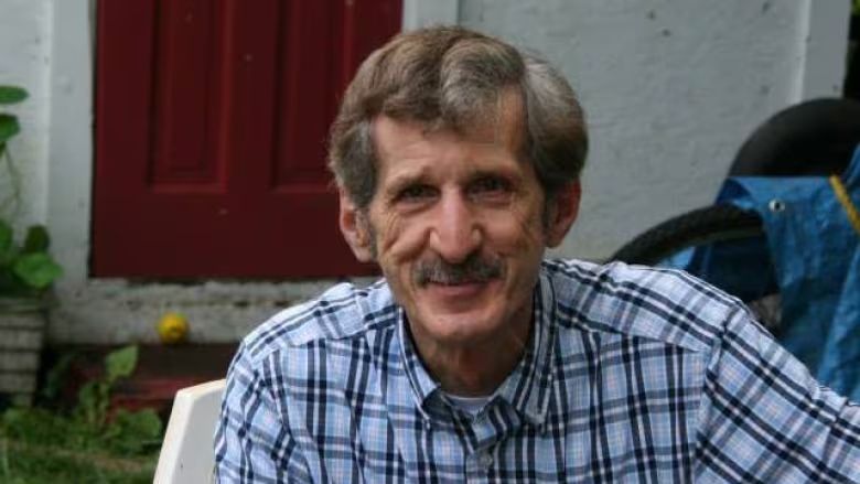 Man with moustache sits in chair and smiles at camera. 