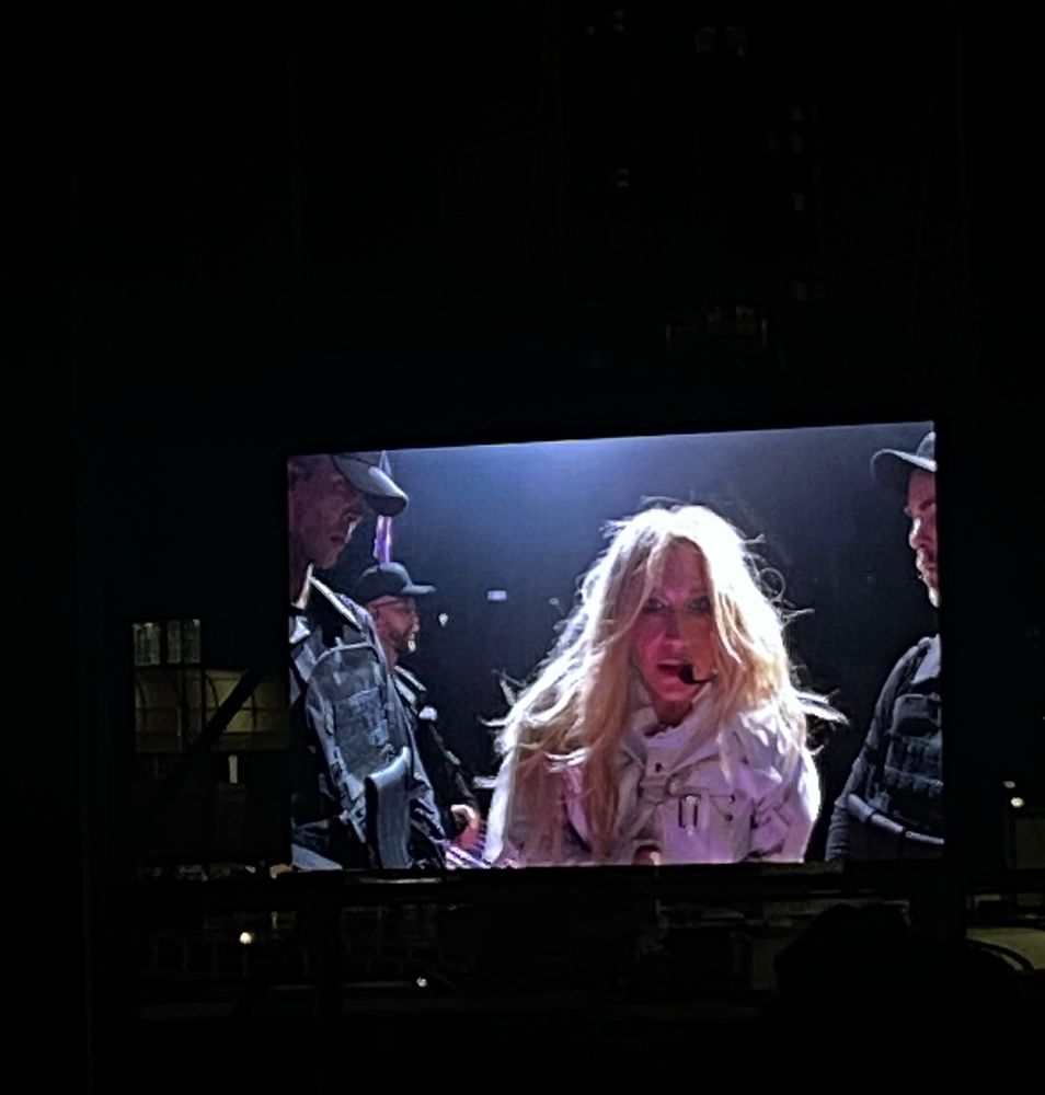A pic of Kesha from the big screen at her concert. She’s mid singing and wearing a white jacket surrounded by guys. 