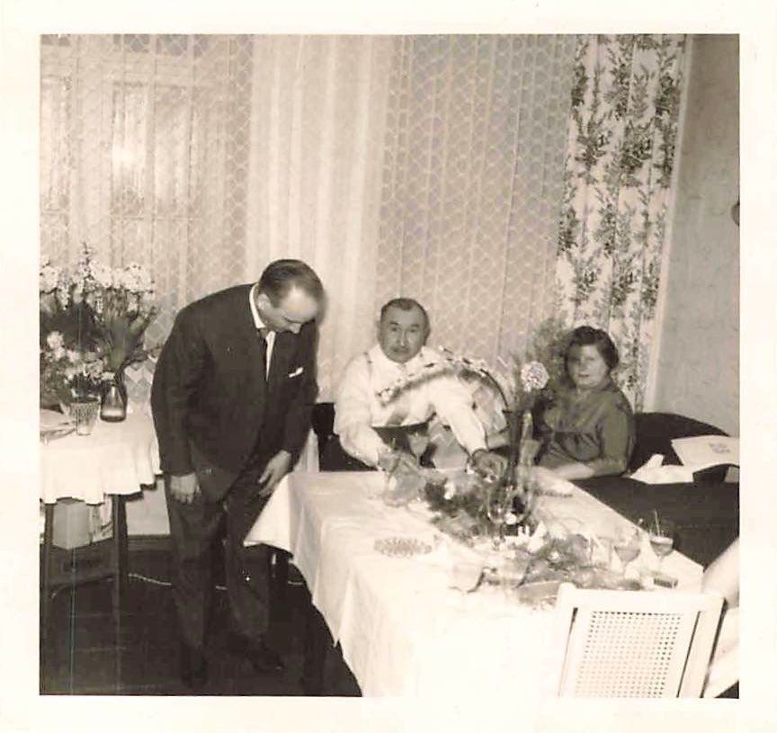 Ein mittelaltes Paar sitzt an einem geschmückten Tisch, ca. 1950er-Jahre. Neben dem Tisch ein Mann, der sich in eine völlig andere Richtung verbeugt.