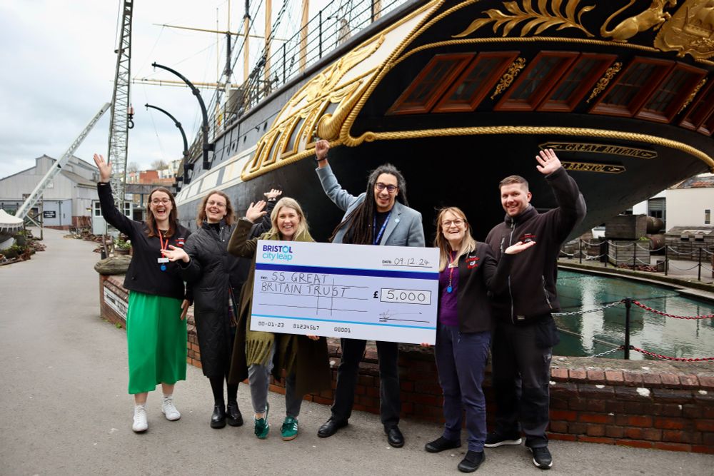 Six individuals stand before the SS Great Britain, proudly holding an oversized cheque for £5,000. The group consists of three Bristol City Leap representatives and three SS Great Britain Trust members, celebrating their collaboration and commitment to community energy initiatives.