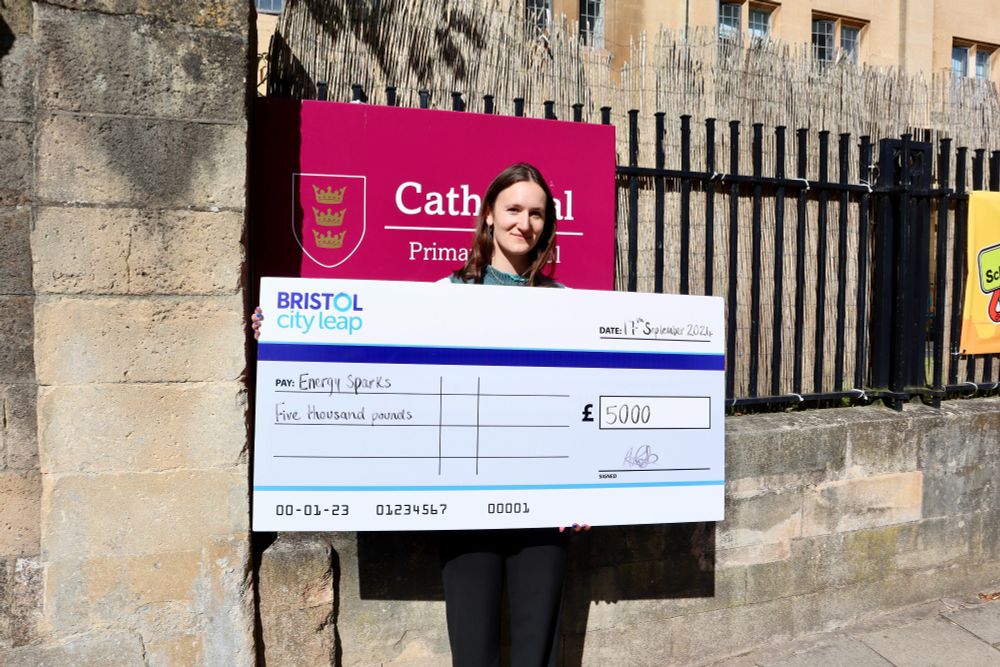 A person stands proudly in front of a school holding a large cheque, celebrating their funding from the Bristol City Leap Community Energy Fund.