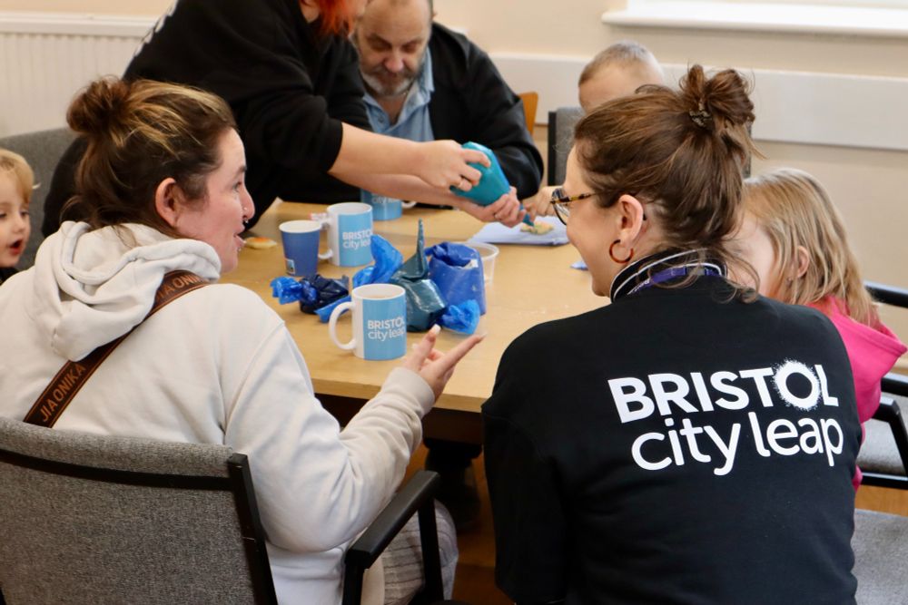 A member of the Bristol City Leap team engages in conversation with a resident at the opening of a newly renovated community room. The event celebrates the completion of a retrofit project designed to enhance the energy efficiency of 81 flats. In the background, several attendees participate in a cheerful biscuit decoration activity.