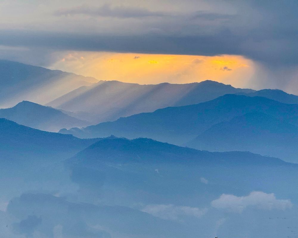 A photo of sunrise over the Himalayas taken from Nagarkot, Nepal on 1 March 2025. The sun's rays appear through a window in thick cloud, gently illuminating the mountains below. The overall tones are blue, apart from the window in the clouds which is pale yellow.