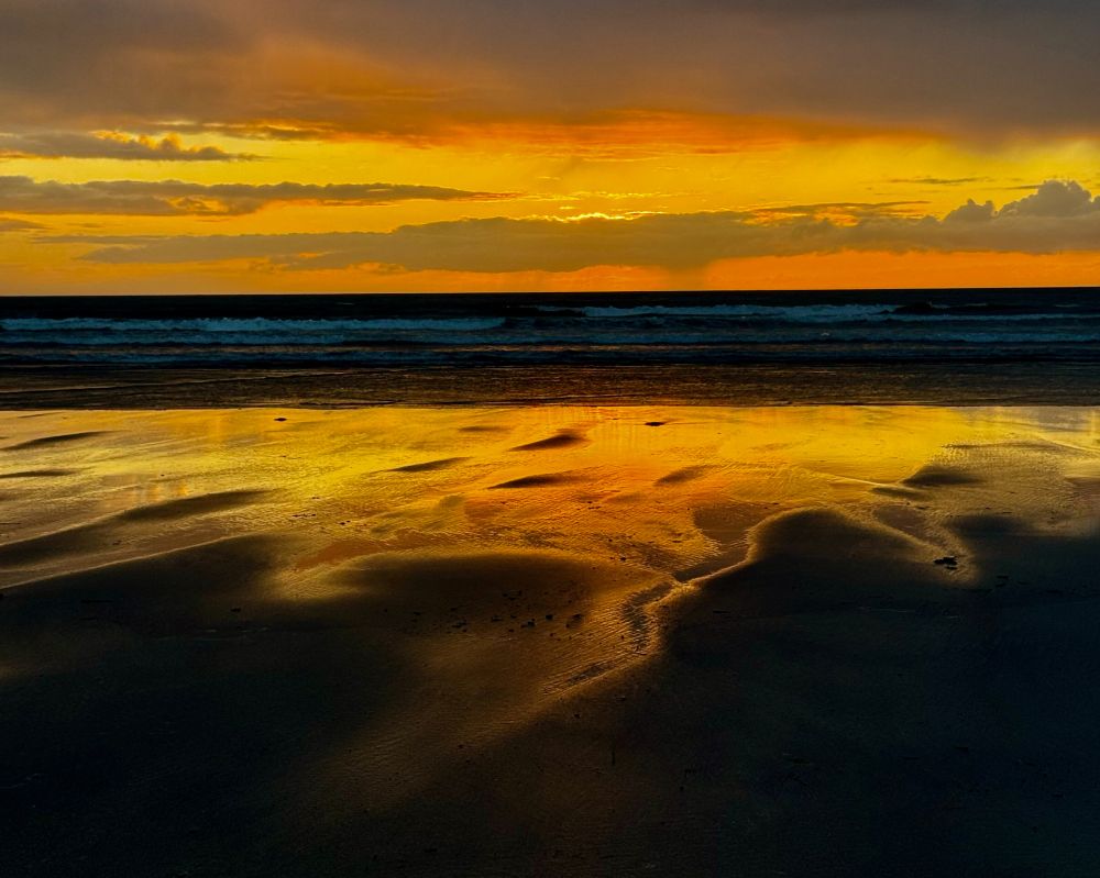 This is a photo of a stunning sundown at Newport Sands, Pembrokeshire, Wales on 25 May 2025. It looks like molten gold has been poured onto the wet sand, with rivulets snaking their way towards the viewer, and contrasting beautifully against the dark tones of the sea and dry sand in the foreground. Although the sun is hidden by clouds, the sky looks like it has been painted with shades of gold. Some white horses on the rolling waves break up the otherwise dark sea which, in turn, provides a dramatic contrast between the golden sky and the golden sand. Even by Newport Sands' high standards, this is a sundown I'll remember for a long time.