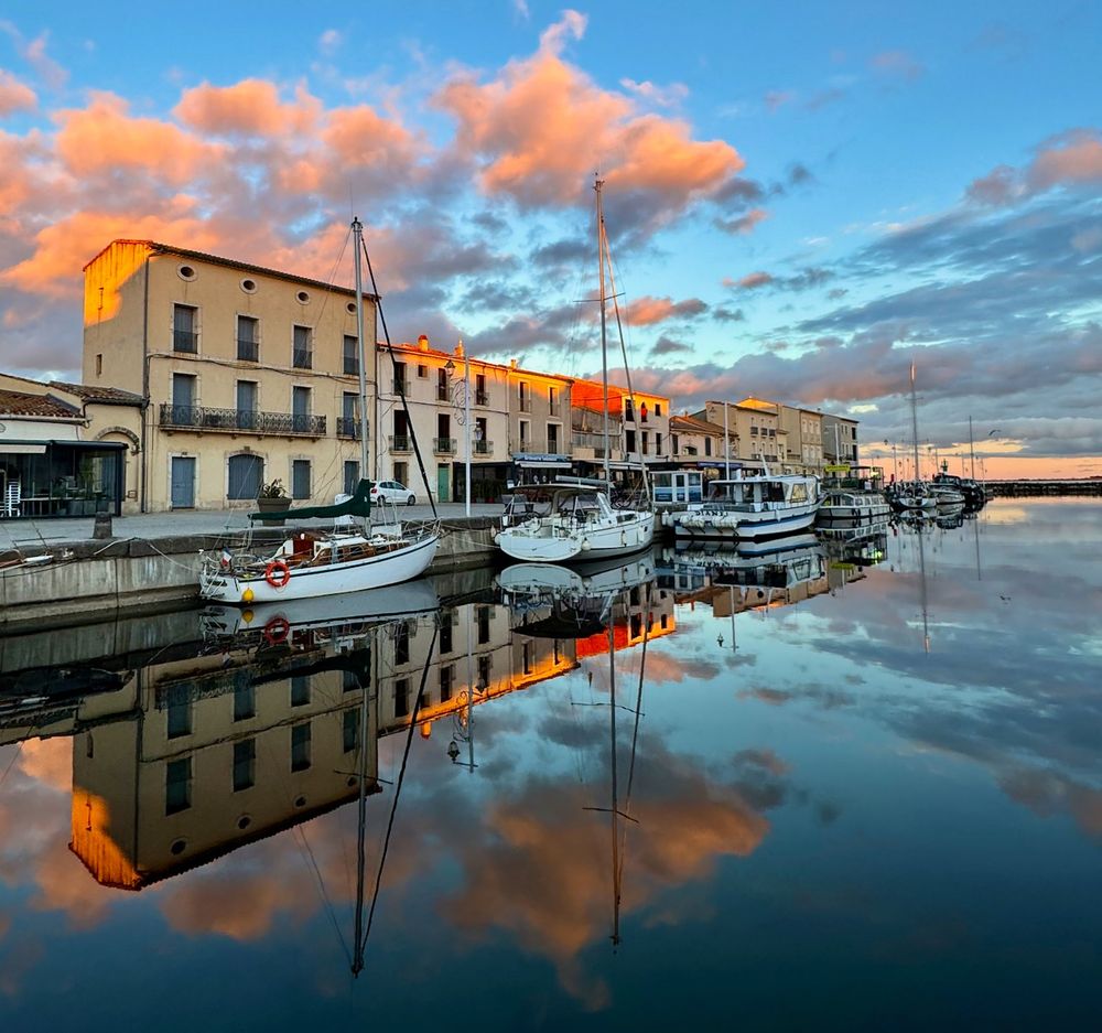 An image of the harbour at Marseillan, Occitanie, France at golden hour in December 2025, with perfect reflections of buildings, boats and pink clouds in the still water. 