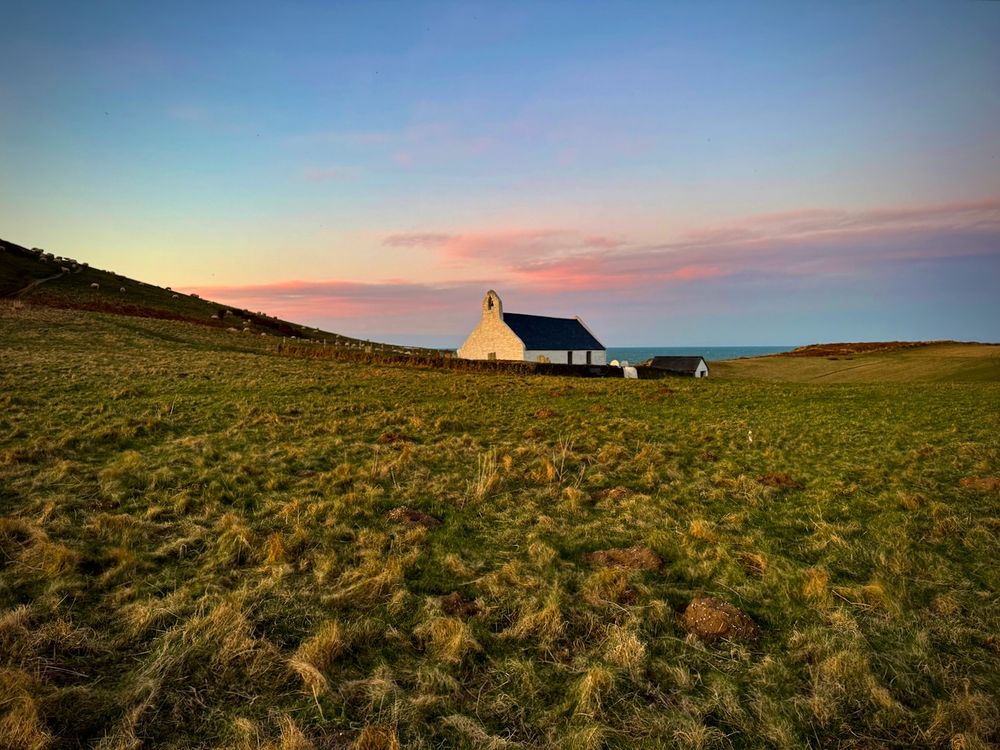 This image shows the beautiful whitewashed Church of the Holy Cross close to the cliffs at Mwnt in Ceredigion, Wales at twilight on 11 March 2025. The sky is pale blue, streaked with pink clouds, and grassy clumps highlighted yellow by the last rays of the sun are in the foreground. The sea can be seen in the distance.
