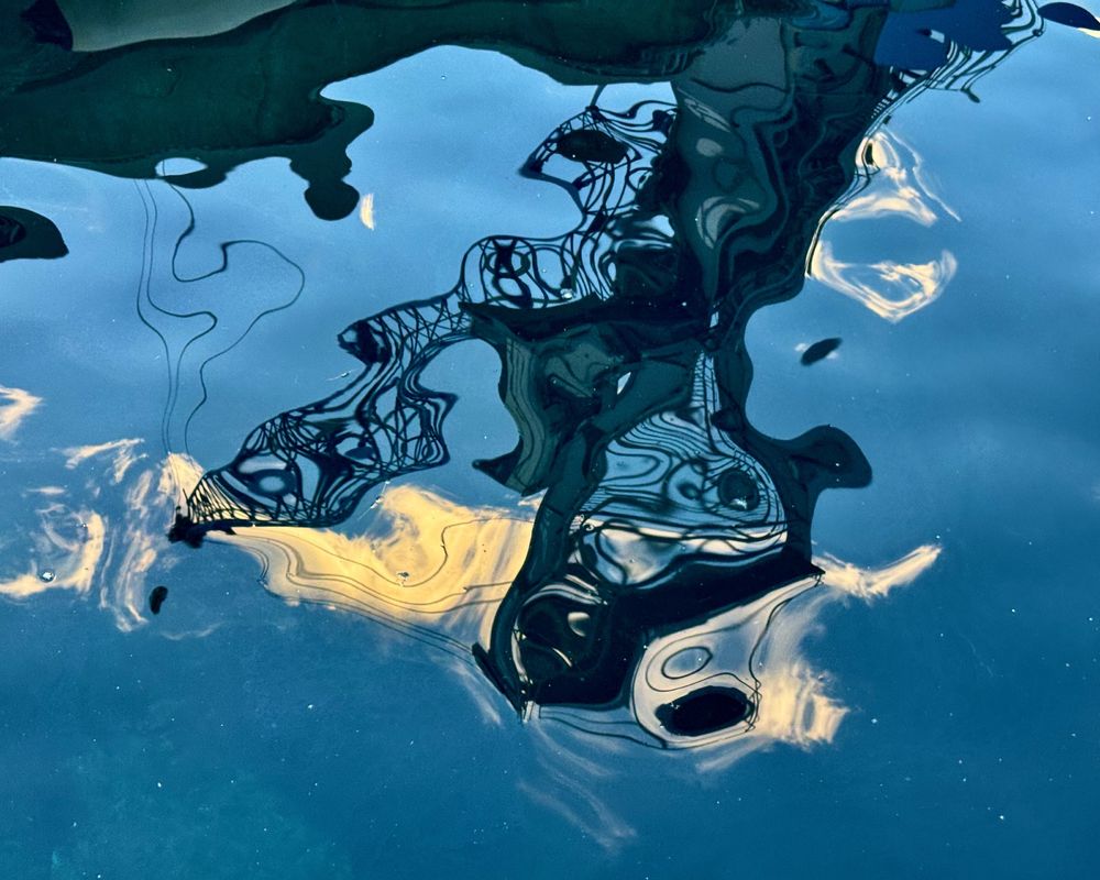 This image shows the reflection of a crane and some wispy clouds in the undulating water of a harbour near Barcelona, Spain. The background is vivid blue, marbled with some darker patches that look almost black. Featuring some Miro-esque elements, the reflection of the crane is a maze of wiggly lines that look as if they have been scribbled on the water.