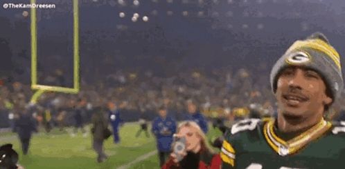 a man wearing a green bay packers jersey and a beanie is standing on a field with his arms in the air .