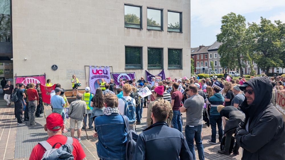 A crowd of people facing a row of UCU banners against a white brick building 