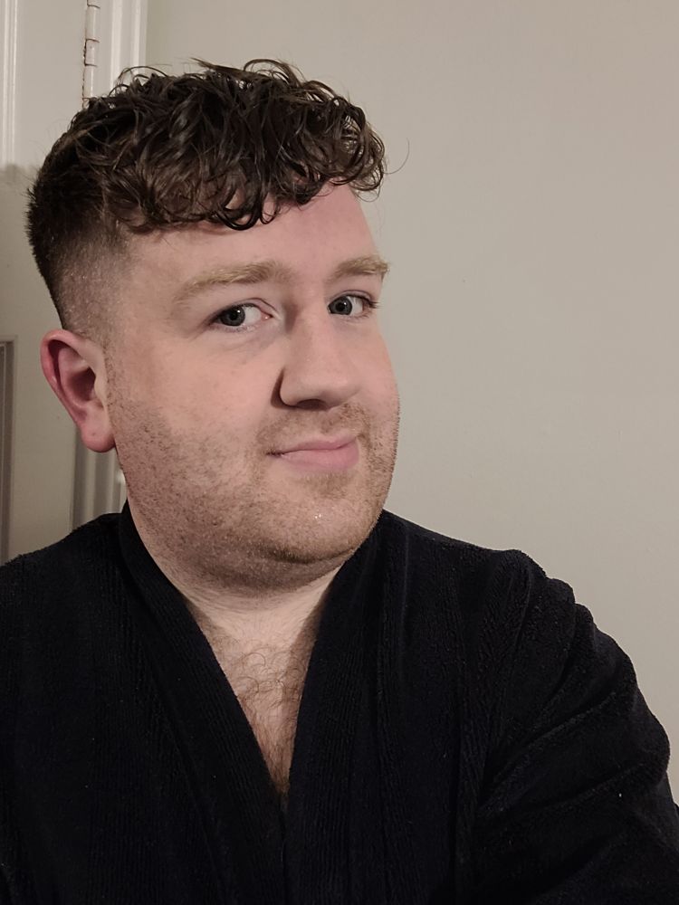 A man in his mid-thirties with wet hair and wearing a navy dressing gown awkwardly takes a selfie